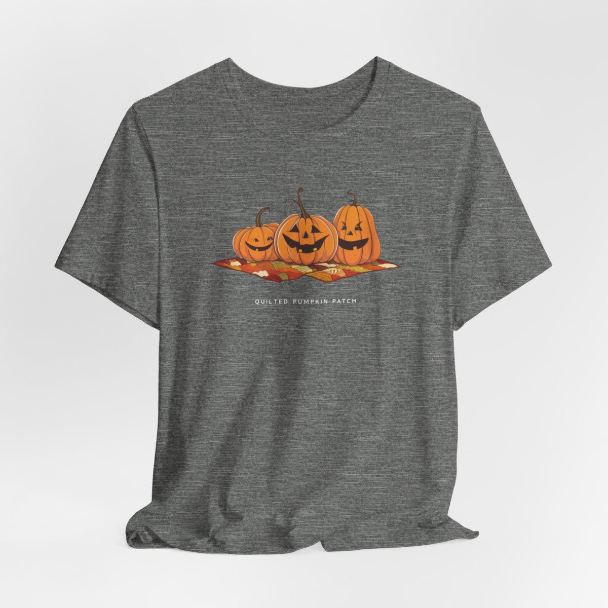 A Deep Heather T-shirt with three smiling jack-o'-lanterns sitting on a colorful autumn-themed quilt. The pumpkins have carved faces with various expressions, and the quilt beneath them features a patchwork of fall colors. Text below reads "QUILTED PUMPKIN PATCH".