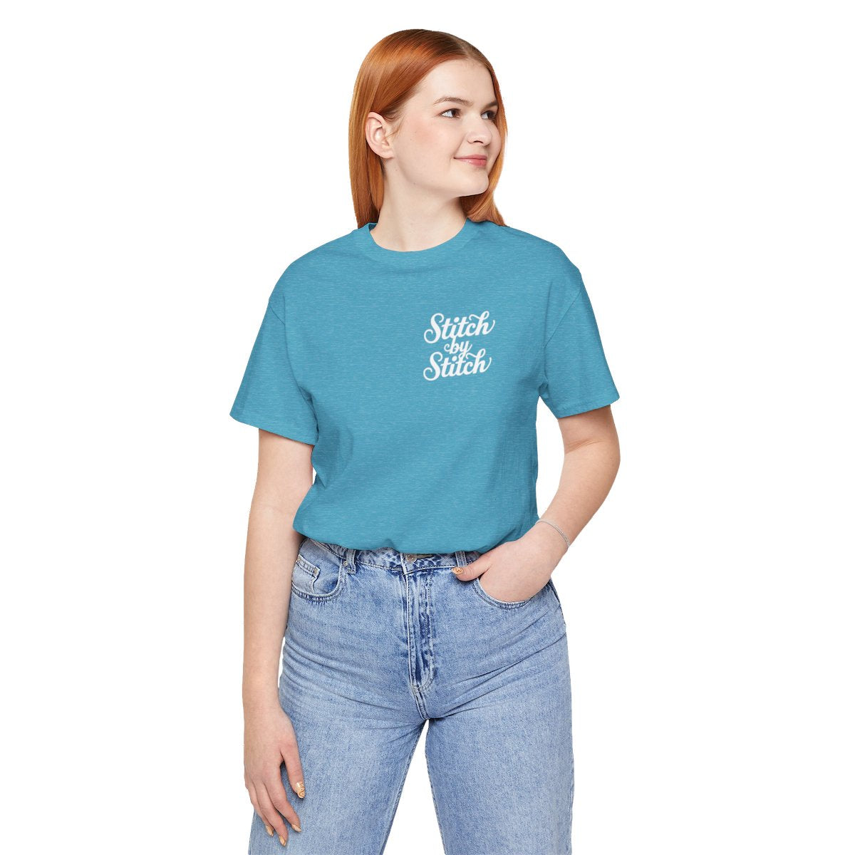 A Heather Aqua T-Shirt featuring the phrase "Stitch by Stitch". The word "Stitch" is written twice in a flowing script font, while "by" is centered between them in a smaller, sans-serif font. The design has a clean, minimalist aesthetic.