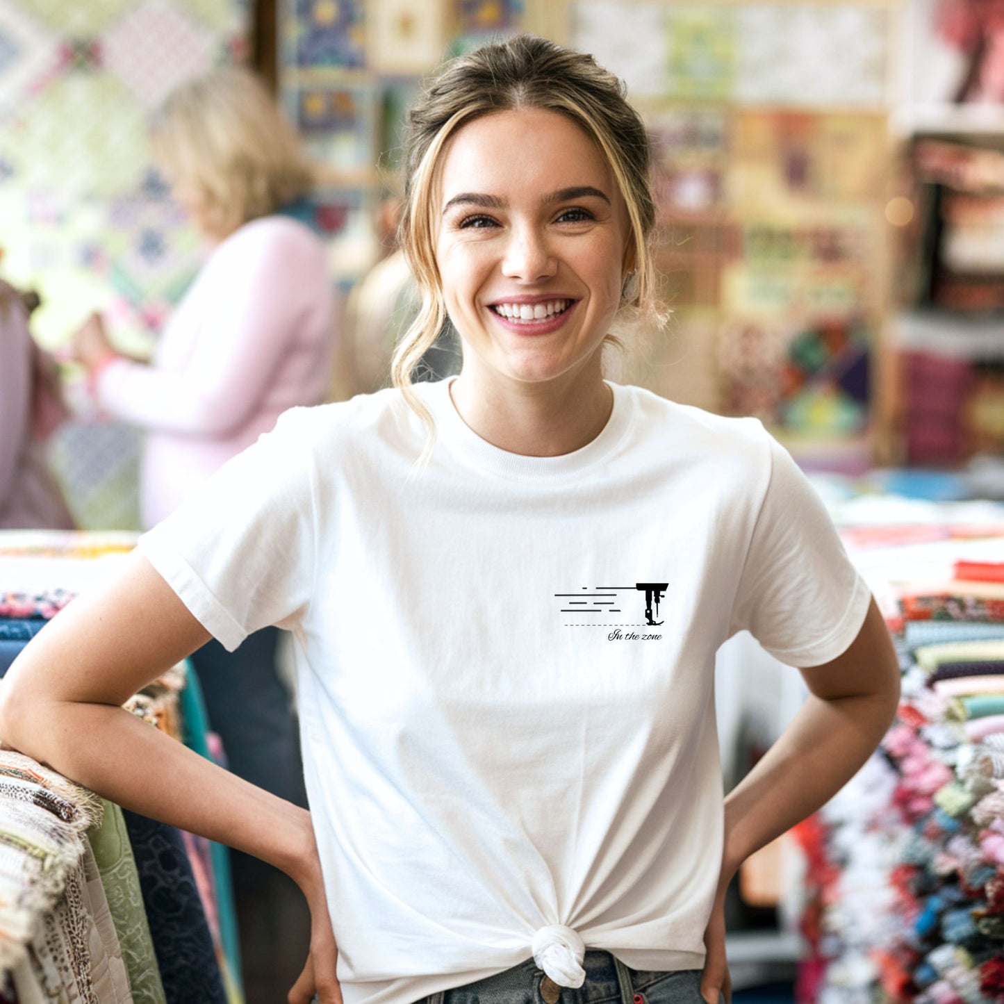 A T-shirt featuring a minimalist black silhouette of a sewing machine arm with motion lines. Below, the phrase "In the zone" is written in cursive script. The design conveys the focus and rhythm of quilting.