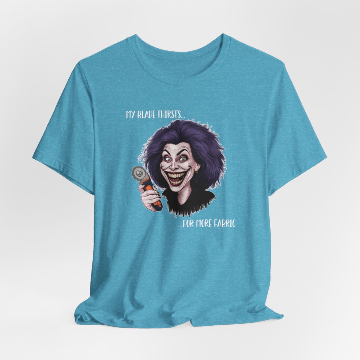 A Heather Aqua T-shirt showing a cartoonish character with wild purple hair and an exaggerated grin, holding a rotary cutter. Text reads "My blade thirsts... for more fabric", humorously depicting a quilter's endless desire for new materials.