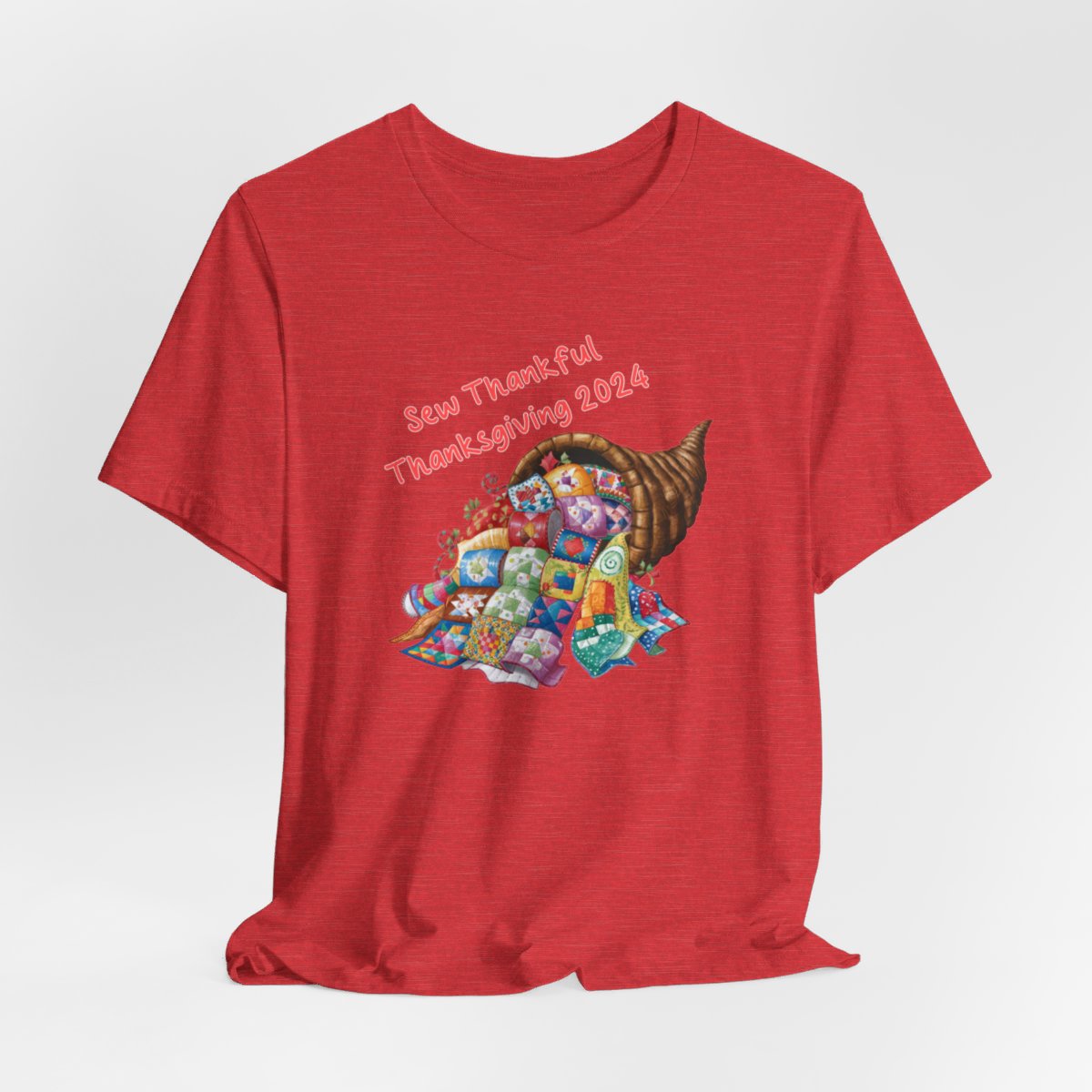 A Heather Red thanksgiving quilting t-shirt featuring a colorful quilted cornucopia overflowing with various quilt patterns and designs. Text reads "Sew Thankful Thanksgiving 2024" in red. Vibrant illustration combines quilting motifs with Thanksgiving imagery.
