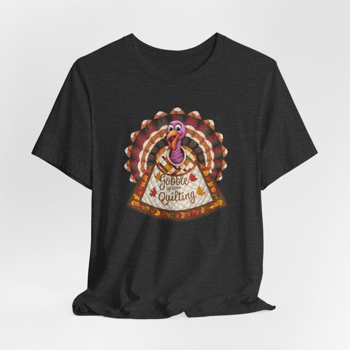 A Dark Grey Heather thanksgiving quilting t-shirt featuring a quilted turkey design with colorful patchwork feathers and body. Text on turkey reads "Gobble up some Quilting" surrounded by fall leaves. Combines Thanksgiving imagery with quilting motifs in a whimsical illustration.