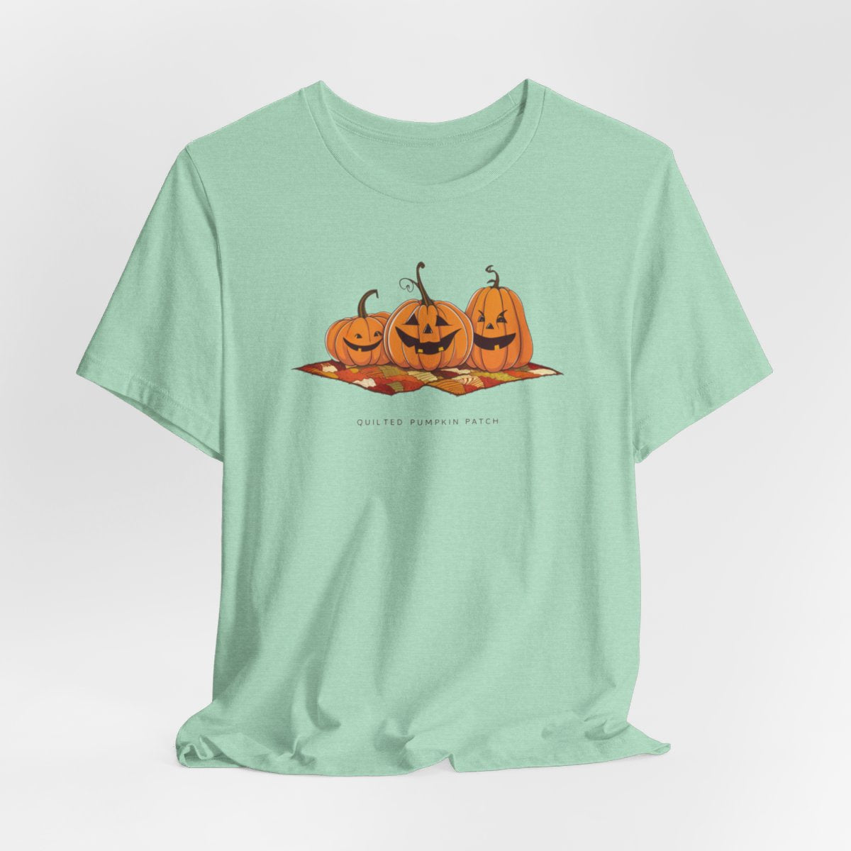 A Heather Mint T-shirt with three smiling jack-o'-lanterns sitting on a colorful autumn-themed quilt. The pumpkins have carved faces with various expressions, and the quilt beneath them features a patchwork of fall colors. Text below reads "QUILTED PUMPKIN PATCH".