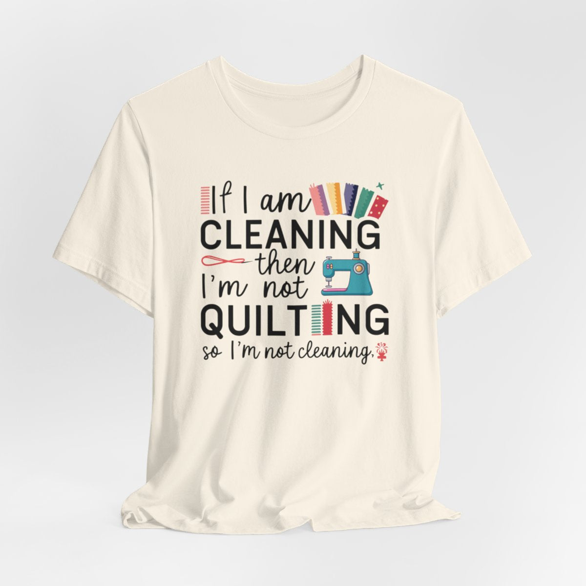 A Natural funny quilting T-Shirt with the phrase If I am Cleaning then I'm not Quilting in a bold and cheerful font with images of quilting icons and supplies