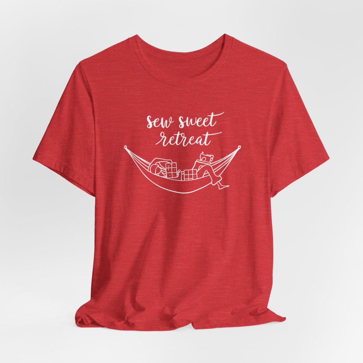 A Heather Red T-Shirt with a black and white illustration of a hammock filled with quilting supplies like fabric squares and a sewing machine. Text above reads "Sew Sweet Retreat" in a flowing script font.