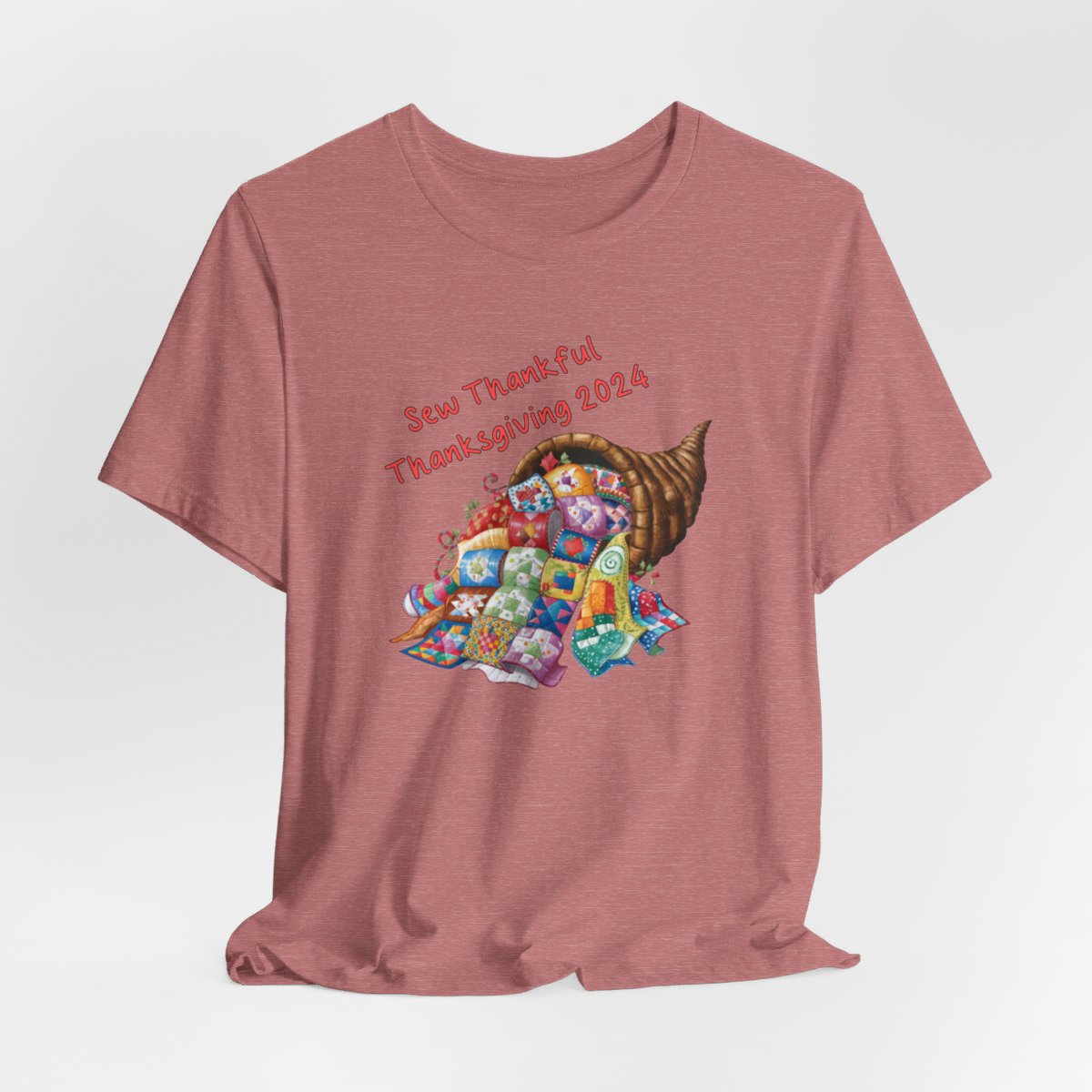 A Heather Mauve thanksgiving quilting t-shirt featuring a colorful quilted cornucopia overflowing with various quilt patterns and designs. Text reads "Sew Thankful Thanksgiving 2024" in red. Vibrant illustration combines quilting motifs with Thanksgiving imagery.