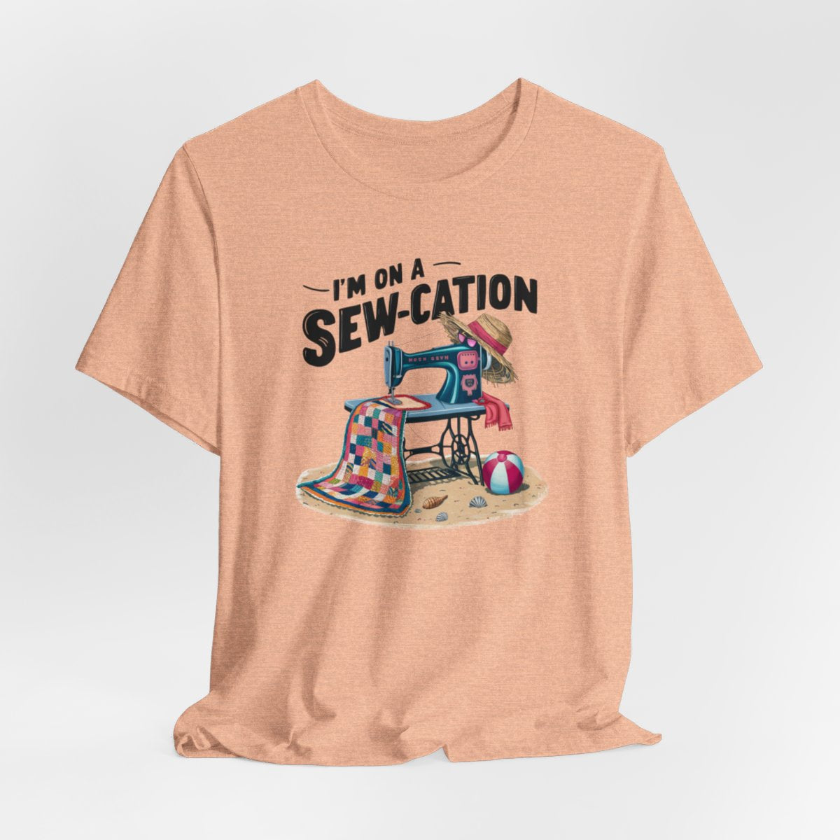A Heather Peach fun Quilting T-shirt of a vintage sewing machine on a beach with a colorful quilt, sun hat, and beach ball. Text above reads "I'm on a Sew-cation" in bold letters.