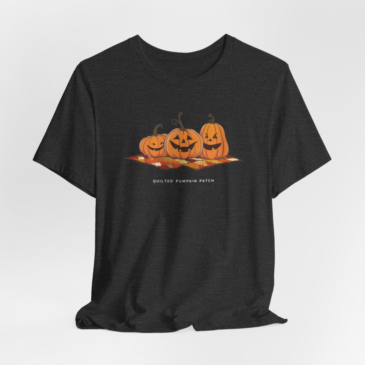 A Dark Grey Heather T-shirt with three smiling jack-o'-lanterns sitting on a colorful autumn-themed quilt. The pumpkins have carved faces with various expressions, and the quilt beneath them features a patchwork of fall colors. Text below reads "QUILTED PUMPKIN PATCH".