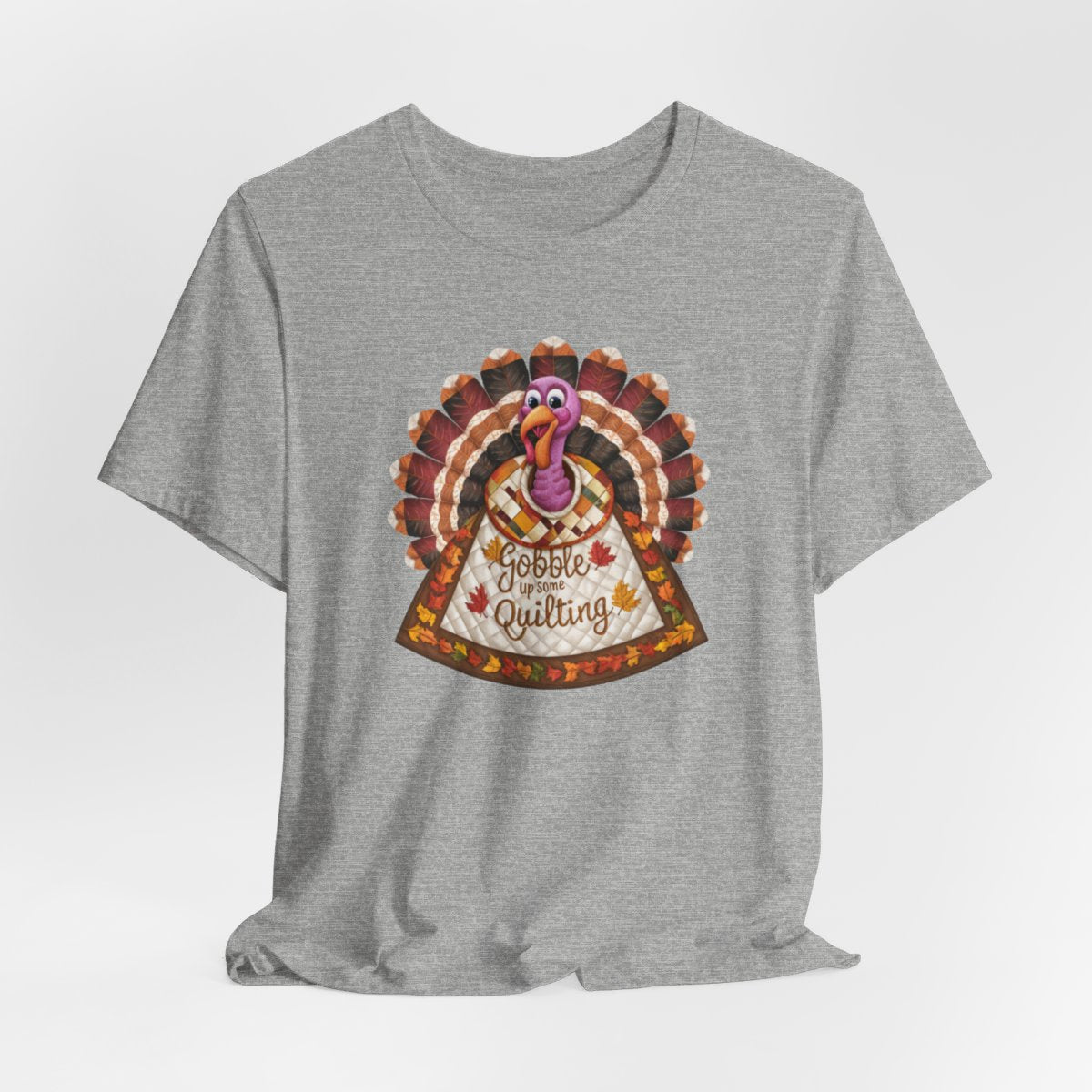 An Athletic Heather thanksgiving quilting t-shirt featuring a quilted turkey design with colorful patchwork feathers and body. Text on turkey reads "Gobble up some Quilting" surrounded by fall leaves. Combines Thanksgiving imagery with quilting motifs in a whimsical illustration.