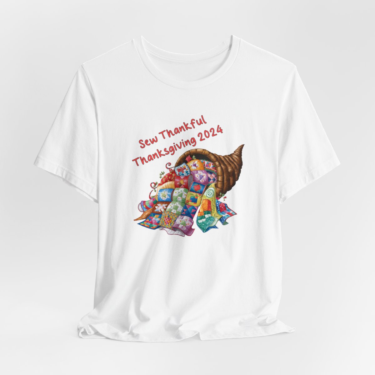 A White thanksgiving quilting t-shirt featuring a colorful quilted cornucopia overflowing with various quilt patterns and designs. Text reads "Sew Thankful Thanksgiving 2024" in red. Vibrant illustration combines quilting motifs with Thanksgiving imagery.