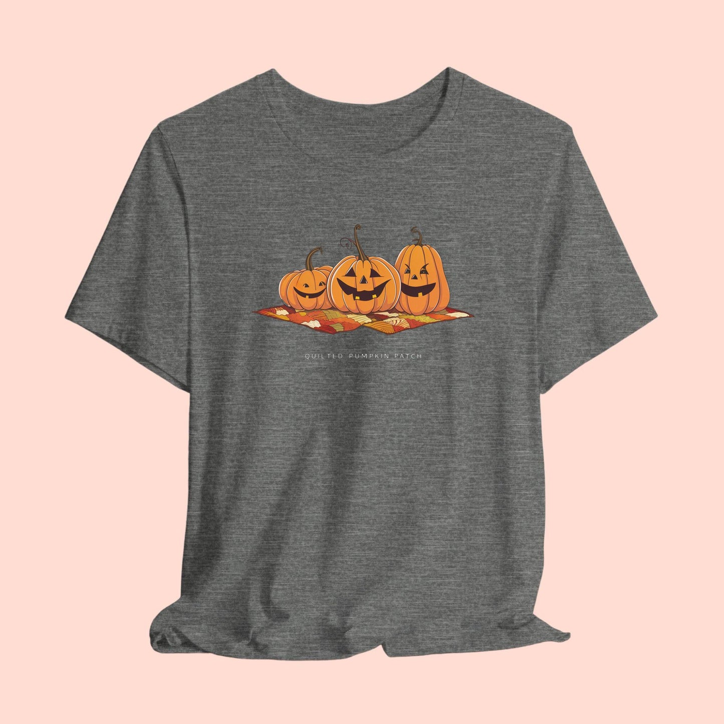 A T-shirt with three smiling jack-o'-lanterns sitting on a colorful autumn-themed quilt. The pumpkins have carved faces with various expressions, and the quilt beneath them features a patchwork of fall colors. Text below reads "QUILTED PUMPKIN PATCH".