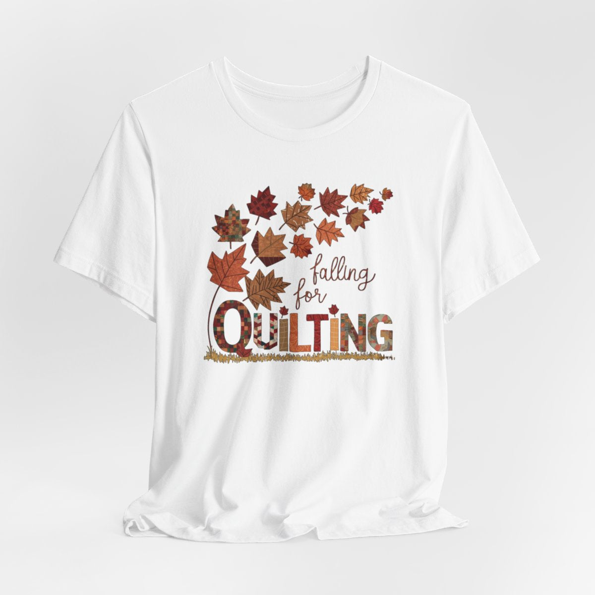 A White thanksgiving quilting t-shirt featuring autumn leaves with various quilting patterns inside. Text reads "falling for QUILTING" with letters in patchwork style. Warm fall colors and quilting motifs combine in a seasonal design.