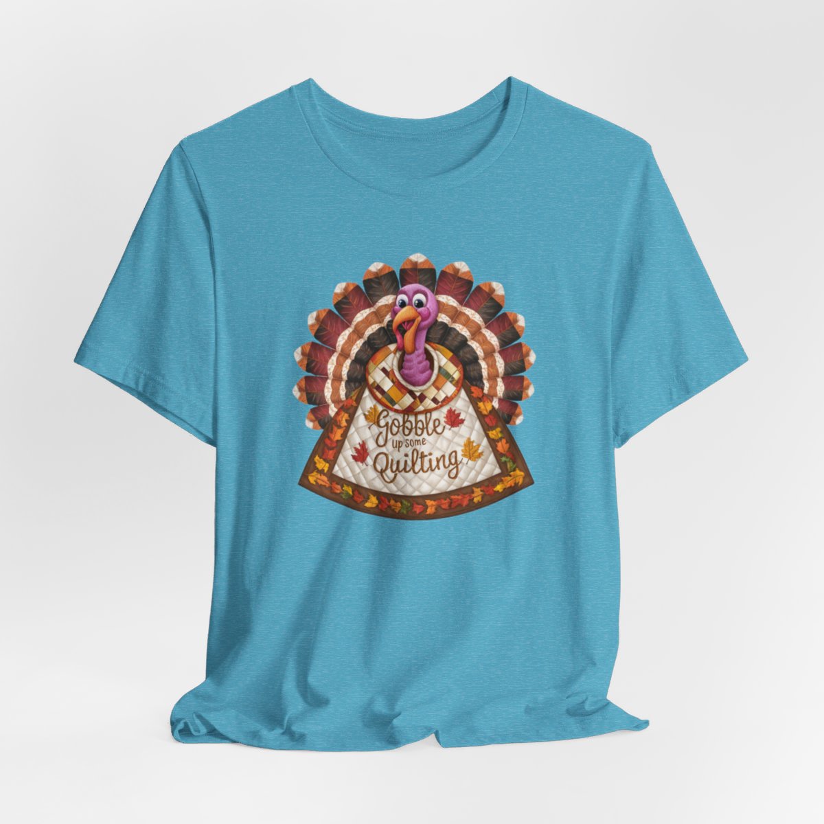 A Heather Aqua thanksgiving quilting t-shirt featuring a quilted turkey design with colorful patchwork feathers and body. Text on turkey reads "Gobble up some Quilting" surrounded by fall leaves. Combines Thanksgiving imagery with quilting motifs in a whimsical illustration.