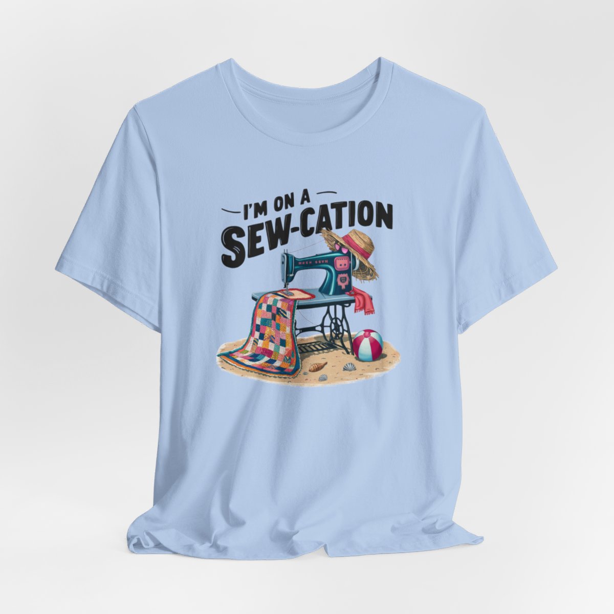 A Baby Blue fun Quilting T-shirt of a vintage sewing machine on a beach with a colorful quilt, sun hat, and beach ball. Text above reads "I'm on a Sew-cation" in bold letters.