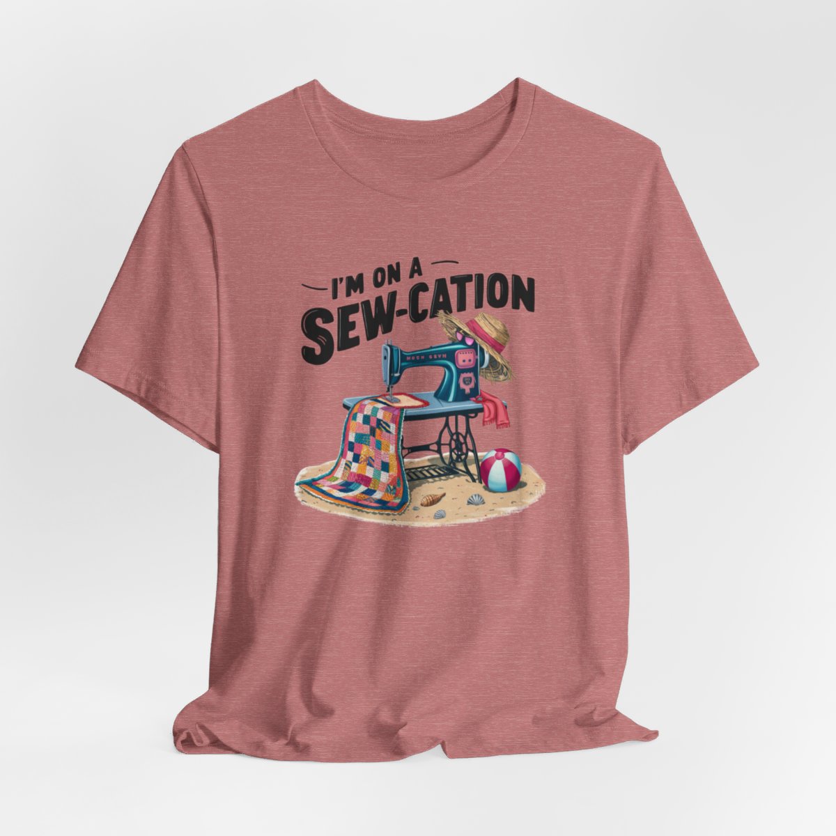 A Heather Mauve fun Quilting T-shirt of a vintage sewing machine on a beach with a colorful quilt, sun hat, and beach ball. Text above reads "I'm on a Sew-cation" in bold letters.