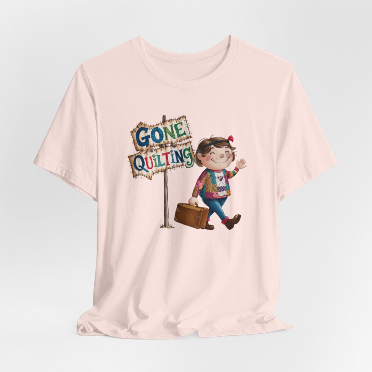A Soft Pinkfun quilting T-Shirt with a cartoon illustration of a smiling girl wearing a patchwork jacket, carrying a suitcase next to a wooden sign that reads "Gone Quilting" in colorful letters.