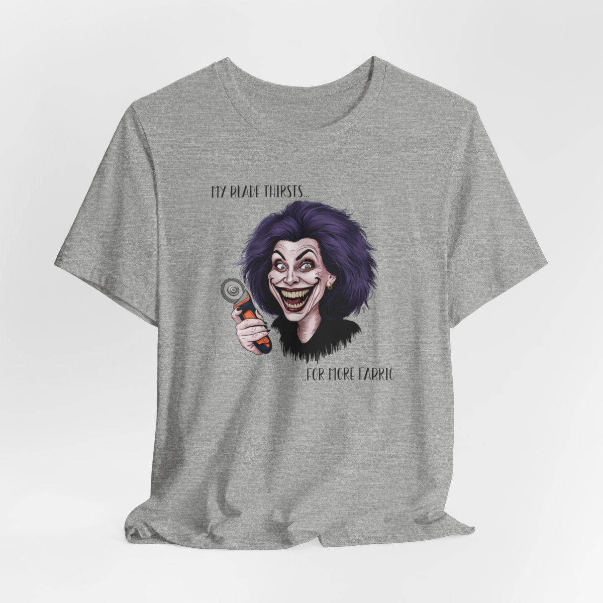 An Athletic Heather T-shirt showing a cartoonish character with wild purple hair and an exaggerated grin, holding a rotary cutter. Text reads "My blade thirsts... for more fabric", humorously depicting a quilter's endless desire for new materials.