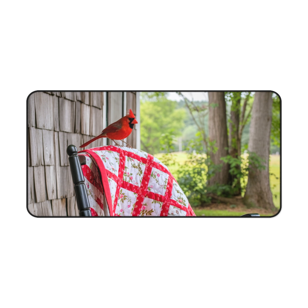 A quilting inspired Desk pad featuring a red cardinal perched on a folded red and white quilt with a floral pattern. The quilt is draped over a wooden chair on a porch, with lush green foliage in the background. Perfect gift for quilters.