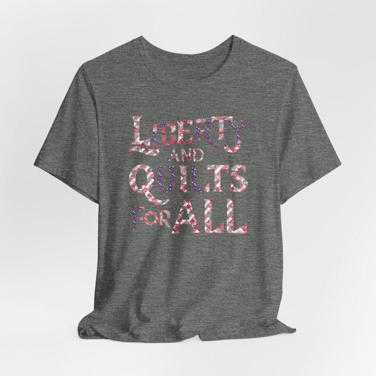 A Deep Heather T-shirt featuring text "LIBERTY AND QUILTS FOR ALL" in large decorative letters filled with various quilt patterns in red, white, and blue.