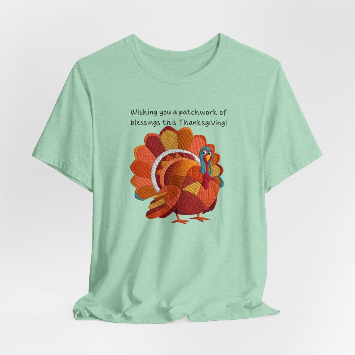 A Heather Mint thanksgiving quilting t-shirt featuring a quilted patchwork turkey in autumn colors with text "Wishing you a patchwork of blessings this Thanksgiving!" Turkey features various quilt patterns and textures in shades of orange, red, yellow, and blue. Whimsical, crafty holiday design perfect for quilting enthusiasts.