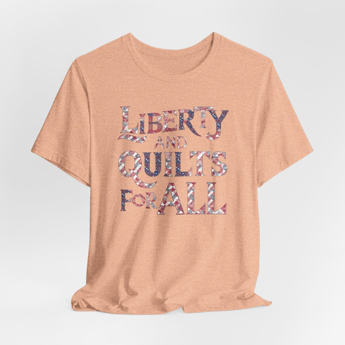 A Heather Peach T-shirt featuring text "LIBERTY AND QUILTS FOR ALL" in large decorative letters filled with various quilt patterns in red, white, and blue.
