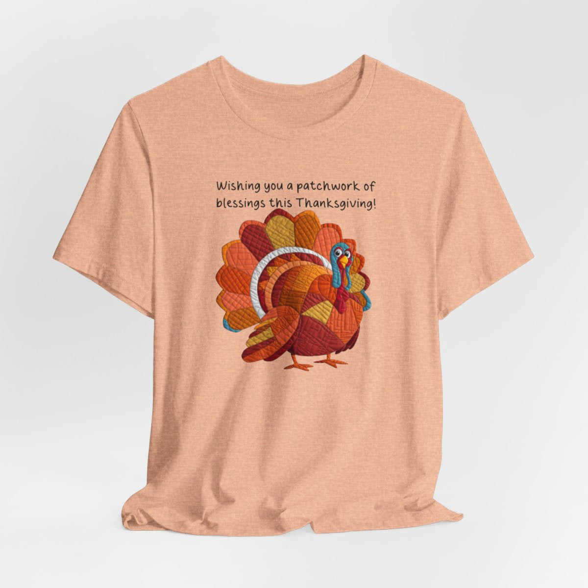 A Heather Peach thanksgiving quilting t-shirt featuring a quilted patchwork turkey in autumn colors with text "Wishing you a patchwork of blessings this Thanksgiving!" Turkey features various quilt patterns and textures in shades of orange, red, yellow, and blue. Whimsical, crafty holiday design perfect for quilting enthusiasts.