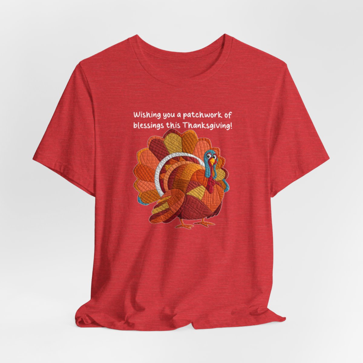 A Heather Red thanksgiving quilting t-shirt featuring a quilted patchwork turkey in autumn colors with text "Wishing you a patchwork of blessings this Thanksgiving!" Turkey features various quilt patterns and textures in shades of orange, red, yellow, and blue. Whimsical, crafty holiday design perfect for quilting enthusiasts.