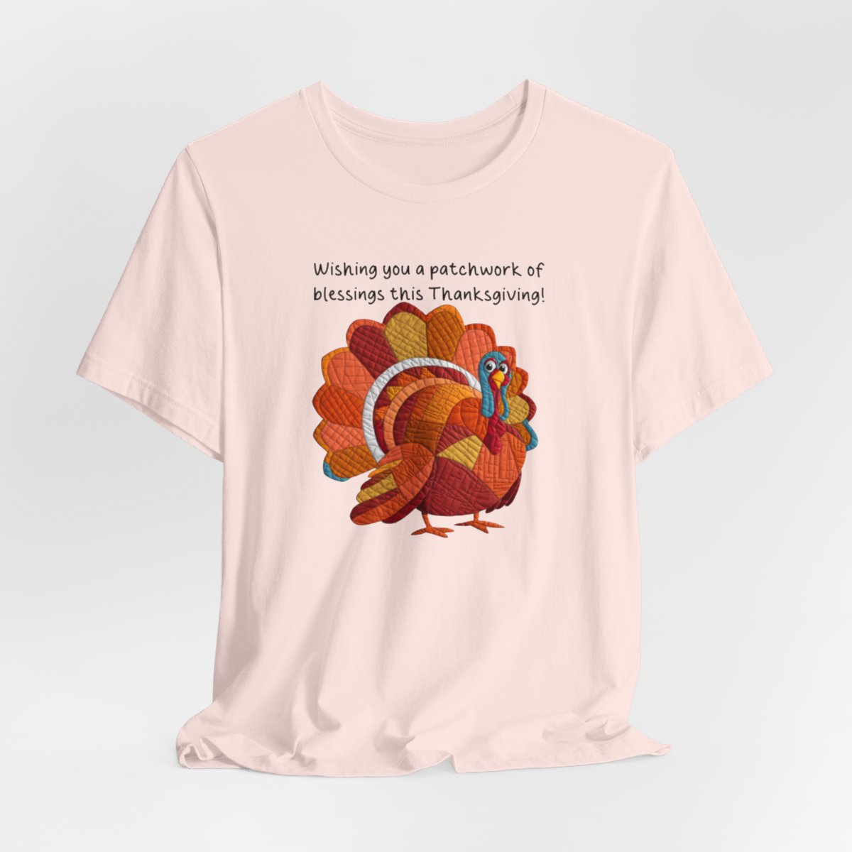 A Soft Pink thanksgiving quilting t-shirt featuring a quilted patchwork turkey in autumn colors with text "Wishing you a patchwork of blessings this Thanksgiving!" Turkey features various quilt patterns and textures in shades of orange, red, yellow, and blue. Whimsical, crafty holiday design perfect for quilting enthusiasts.