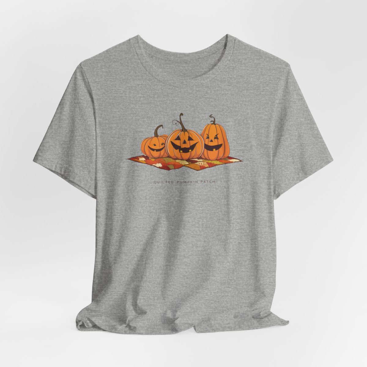 An Athletic Heather T-shirt with three smiling jack-o'-lanterns sitting on a colorful autumn-themed quilt. The pumpkins have carved faces with various expressions, and the quilt beneath them features a patchwork of fall colors. Text below reads "QUILTED PUMPKIN PATCH".