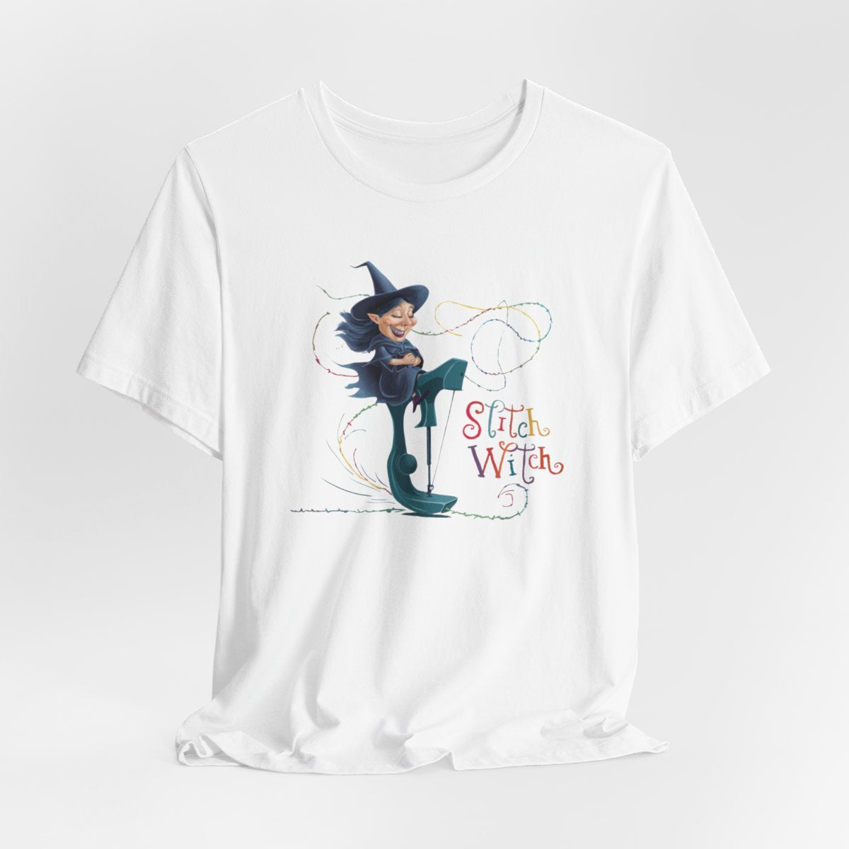 A White T-shirt with a cartoon illustration of a smiling witch with pointed hat riding a teal sewing machine. Colorful stitches swirl around her in the air. Text reads "Stitch Witch" in playful, multicolored letters.