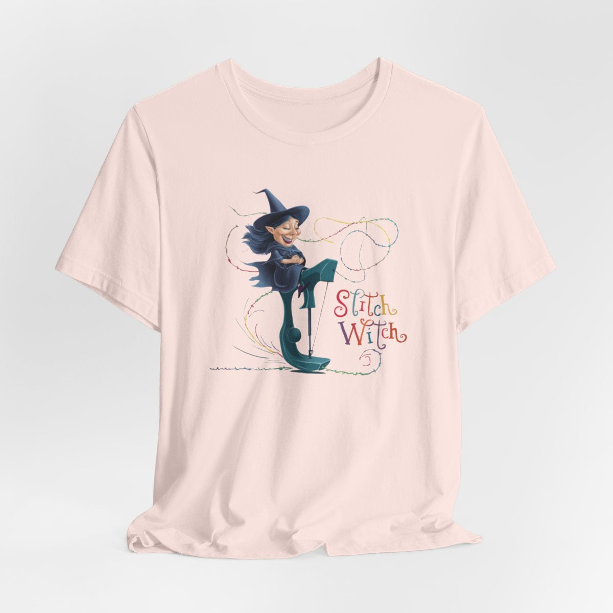A Soft Pink T-shirt with a cartoon illustration of a smiling witch with pointed hat riding a teal sewing machine. Colorful stitches swirl around her in the air. Text reads "Stitch Witch" in playful, multicolored letters.