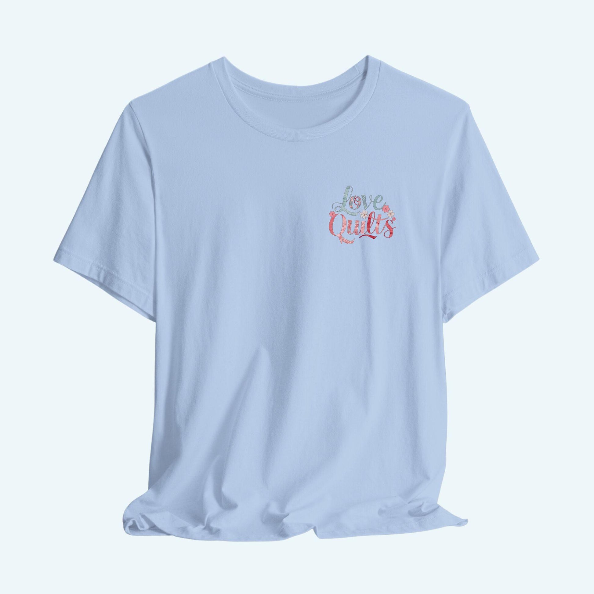 A t-shirt spelling out "Love Quilts". The word "Love" is in teal, while "Quilts" features various shades of pink and red. Small flowers in white and pink adorn the letters, creating a floral and quilting-inspired artwork.