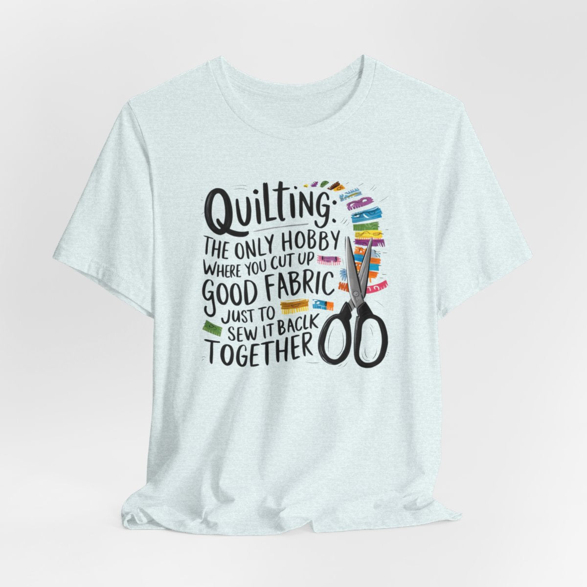A Heather Ice Blue Quilting-themed T-Shirt with text "Quilting: The Only Hobby Where You Cut Up Good Fabric Just to Sew it Back Together" surrounded by colorful fabric scraps and scissors.