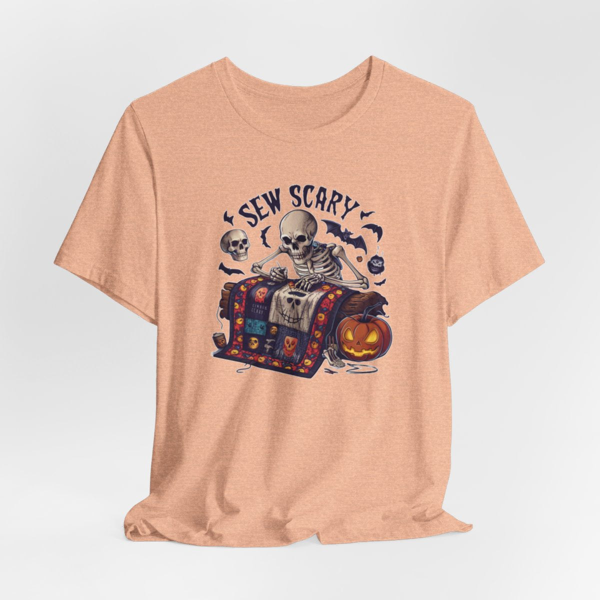 A Heather Peach Halloween Quilting T-Shirt featuring a skeleton sewing Halloween quilt. Colorful patchwork with spooky motifs. Bats and pumpkins surround skeleton. Text reads "Sew Scary". Ideal for quilters who love Halloween.