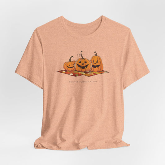 A Heather Peach T-shirt with three smiling jack-o'-lanterns sitting on a colorful autumn-themed quilt. The pumpkins have carved faces with various expressions, and the quilt beneath them features a patchwork of fall colors. Text below reads "QUILTED PUMPKIN PATCH".