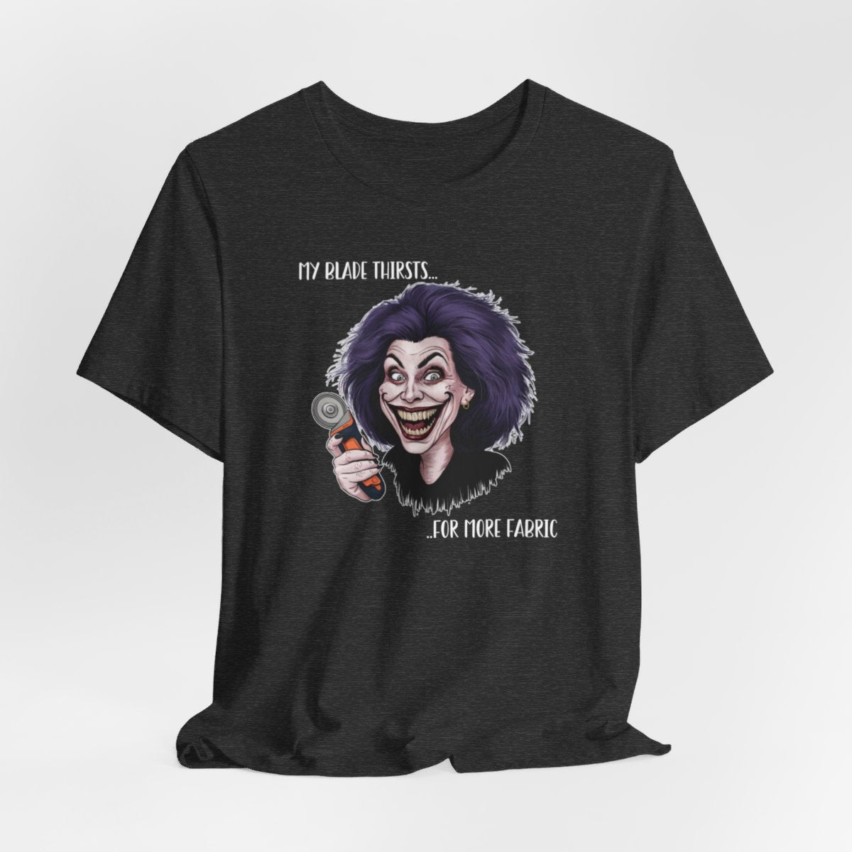 A Dark Grey Heather T-shirt showing a cartoonish character with wild purple hair and an exaggerated grin, holding a rotary cutter. Text reads "My blade thirsts... for more fabric", humorously depicting a quilter's endless desire for new materials.