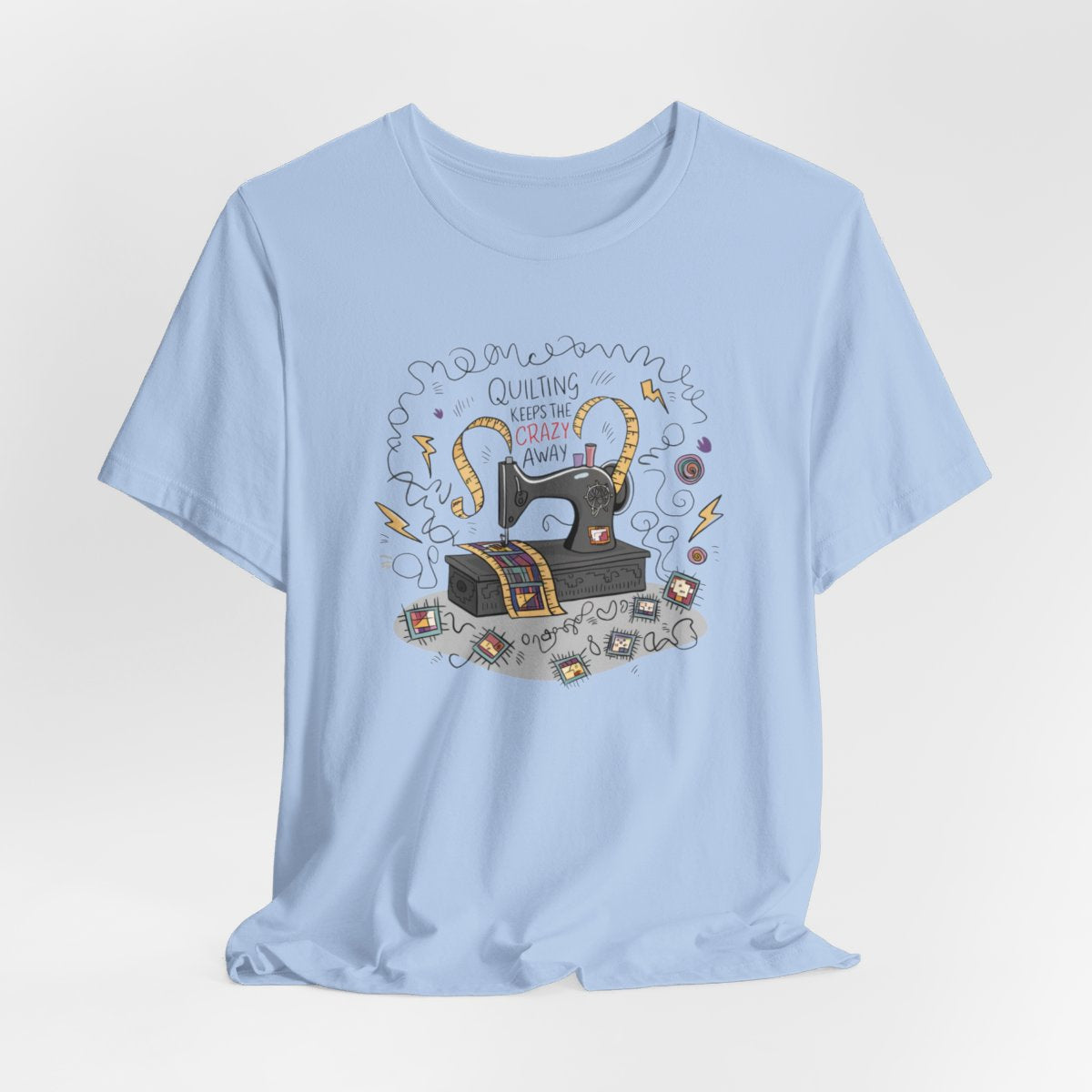 A Baby Blue T-Shirt with a vintage sewing machine, measuring tape heart, and text "Quilting Keeps the Crazy Away" amidst swirls and quilting motifs.