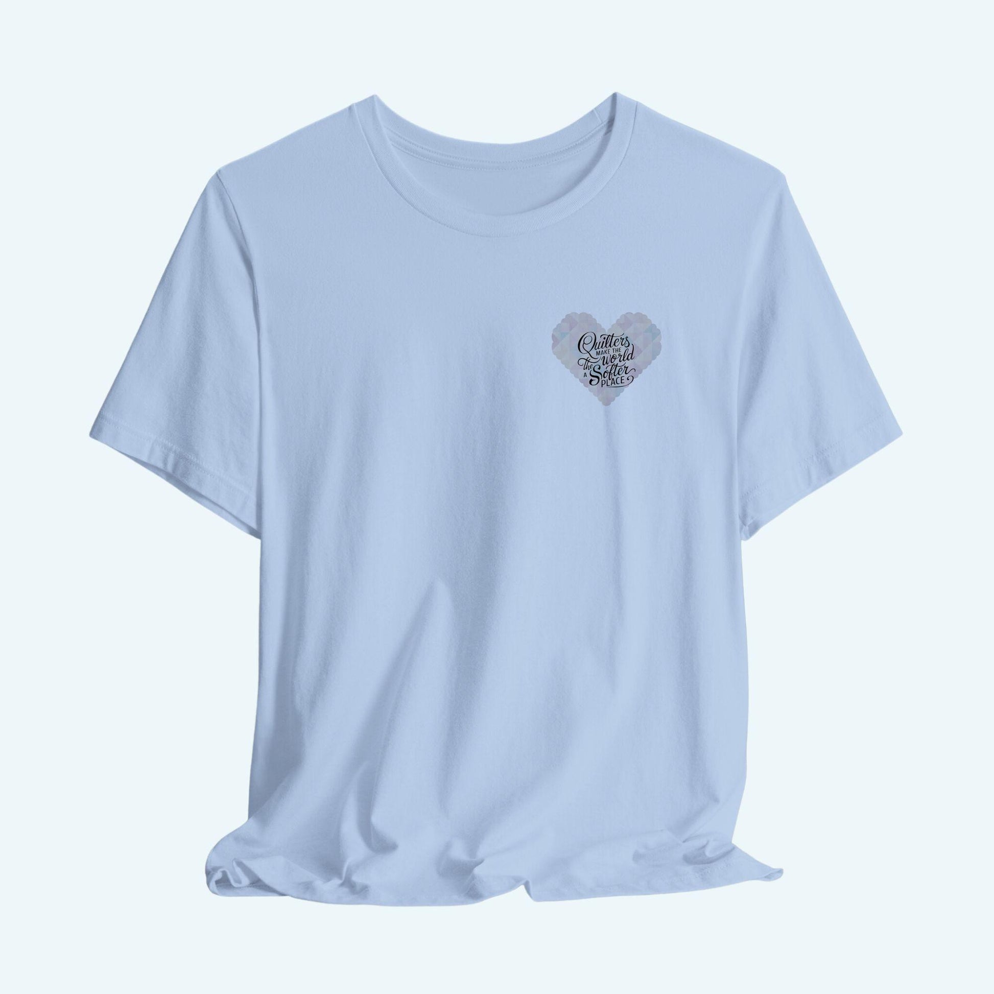 A T-shirt showing a large heart-shaped quilt pattern in pastel colors of pink, blue, and cream. Black text overlaid on the heart reads "Quilters make the world a softer place" in a mix of script and sans-serif fonts. The design combines geometric quilt patterns with an uplifting message about quilting.