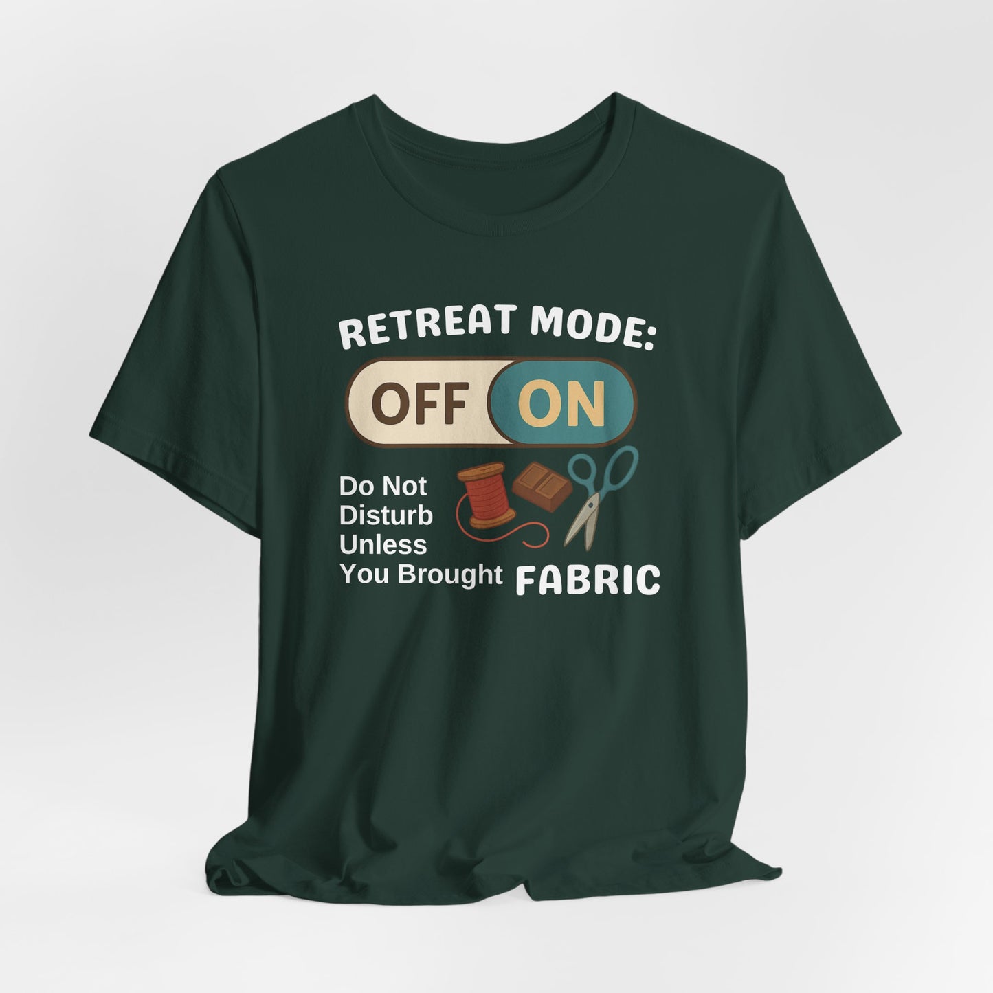 A A Forest t-shirt with a toggle switch that says "Retreat Mode: OFF/ON" surrounded by a spool of thread, chocolate, and scissors. Text below reads "Do not disturb unless you brought fabric." Fun gift for quilting retreats.