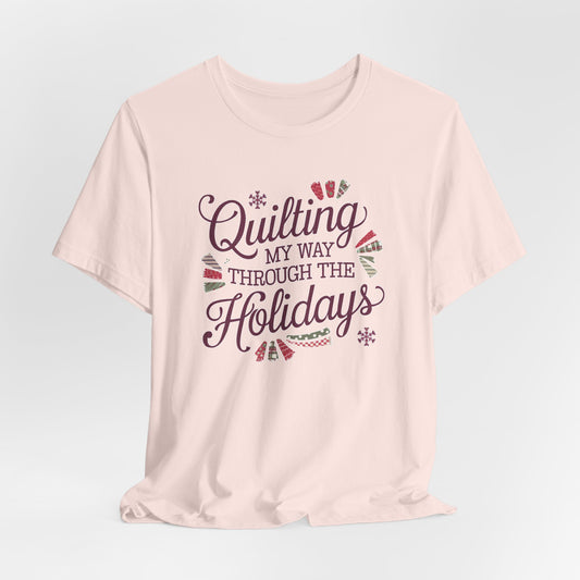 A Soft Pink playful holiday-themed t-shirt with the phrase "Quilting My Way Through the Holidays" surrounded by quilting-inspired decorations like fabric swatches and snowflakes.