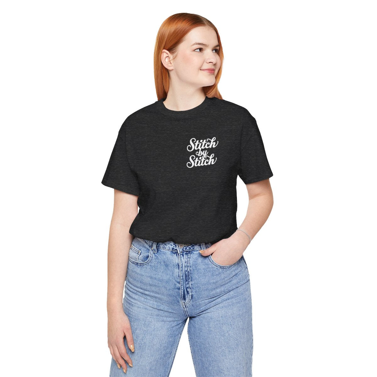 A Dark Grey Heather T-Shirt featuring the phrase "Stitch by Stitch". The word "Stitch" is written twice in a flowing script font, while "by" is centered between them in a smaller, sans-serif font. The design has a clean, minimalist aesthetic.