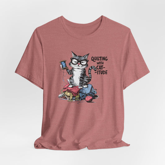 A Heather Mauve t-shirt featuring a sassy cartoon cat sitting on a pile of colorful fabric, holding a spool of thread, with the phrase 'Quilting with Cat-itude,' perfect for quilters and cat lovers.