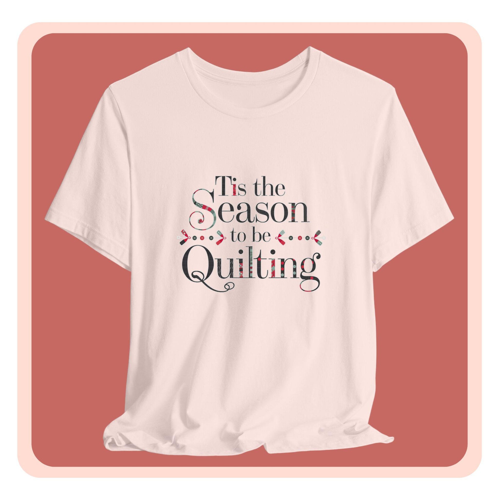 A festive t-shirt featuring the phrase "'Tis the Season to be Quilting," with playful sewing-inspired elements like needles, buttons, and fabric patterns integrated into the typography.