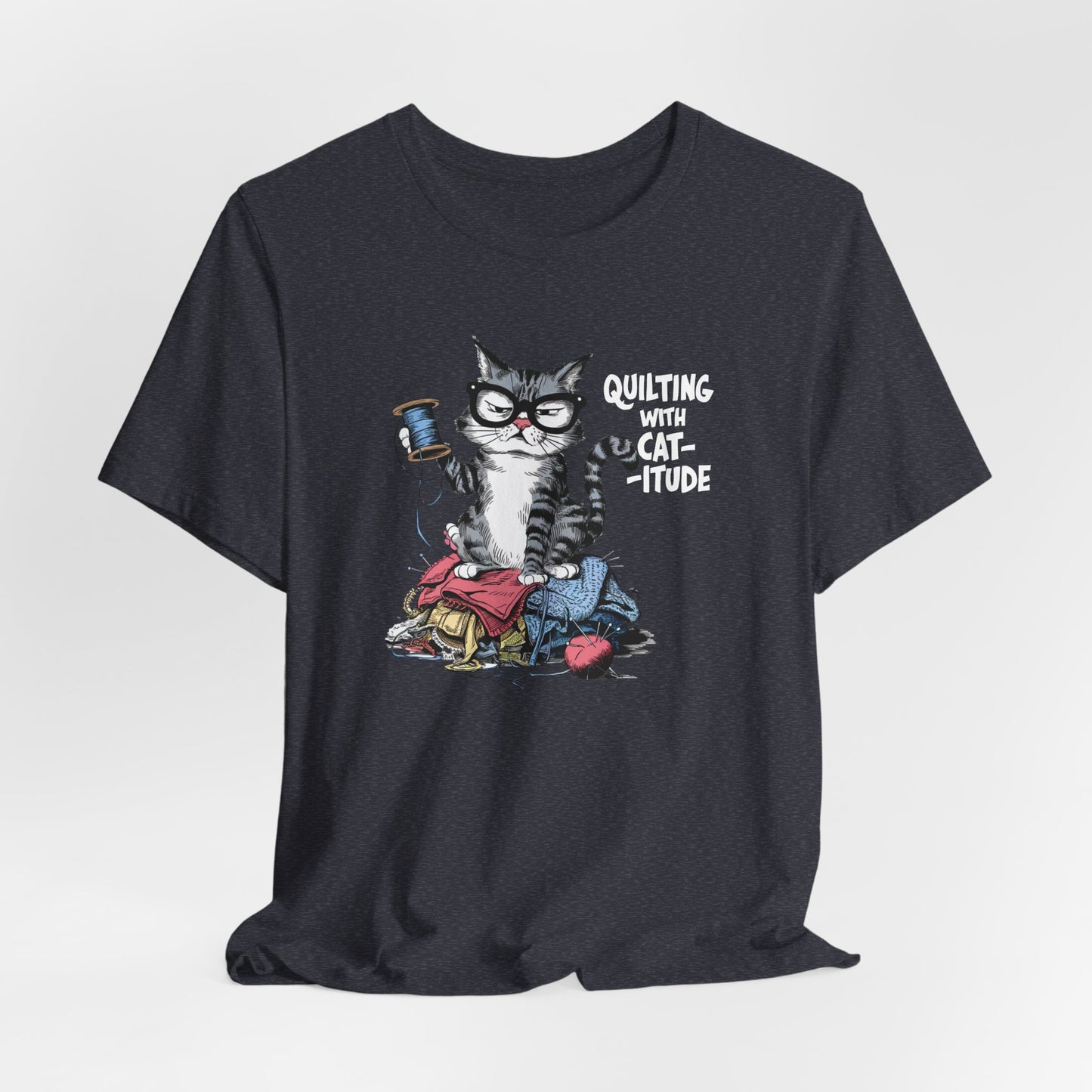 A Heather Navy t-shirt featuring a sassy cartoon cat sitting on a pile of colorful fabric, holding a spool of thread, with the phrase 'Quilting with Cat-itude,' perfect for quilters and cat lovers.