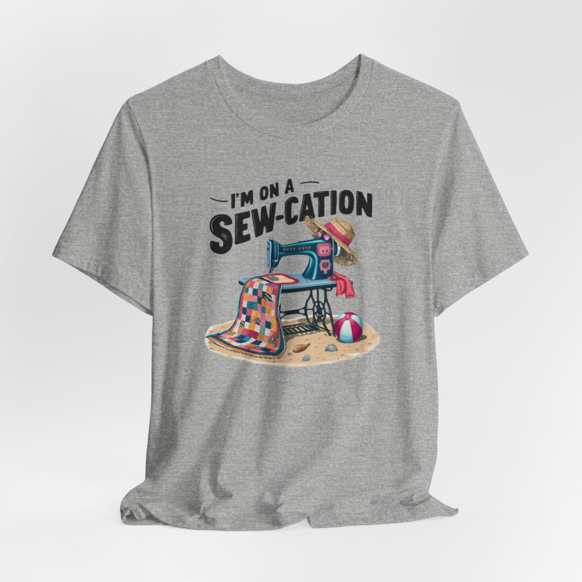 An Athletic Heather fun Quilting T-shirt of a vintage sewing machine on a beach with a colorful quilt, sun hat, and beach ball. Text above reads "I'm on a Sew-cation" in bold letters.