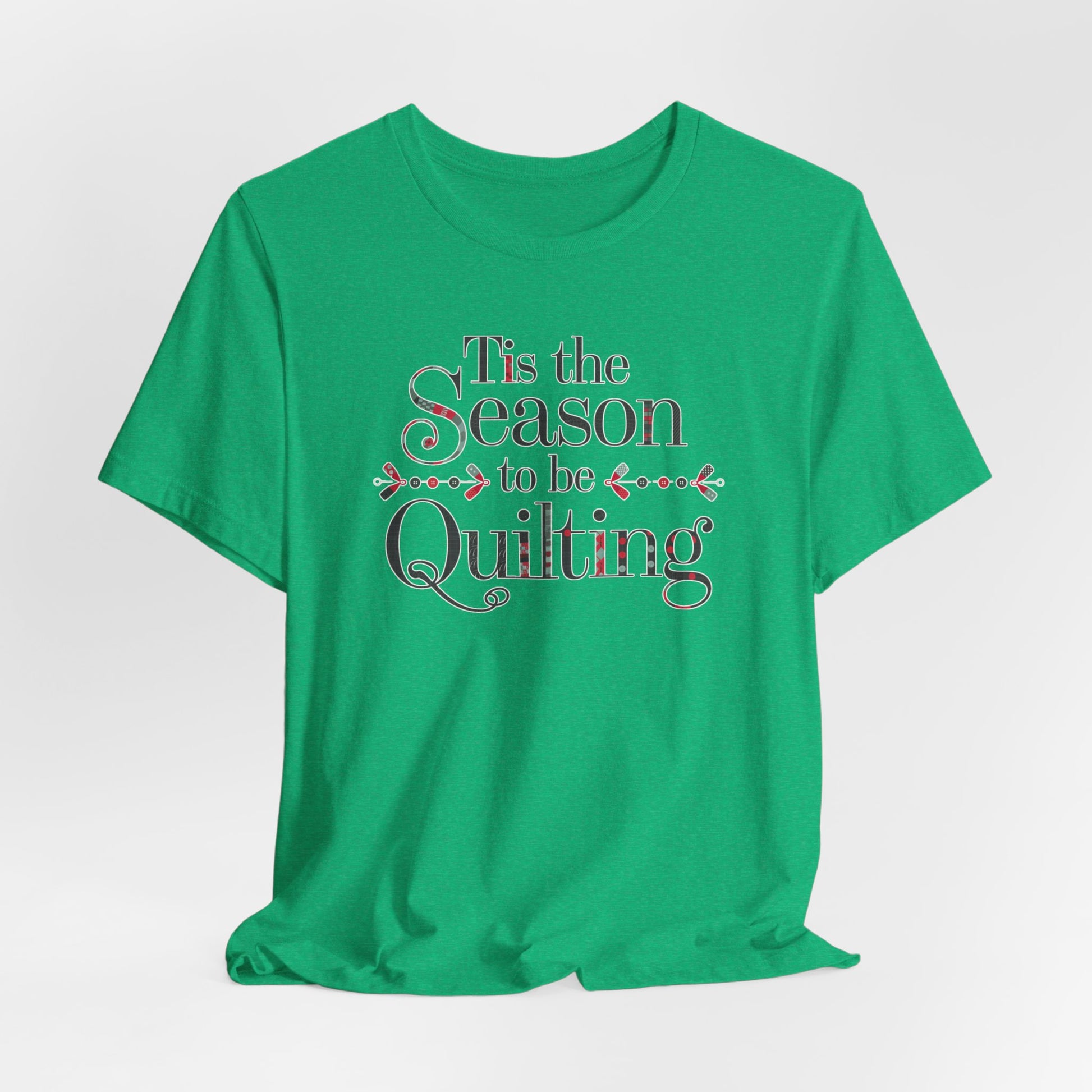 A Heather Kelly festive t-shirt featuring the phrase "'Tis the Season to be Quilting," with playful sewing-inspired elements like needles, buttons, and fabric patterns integrated into the typography.