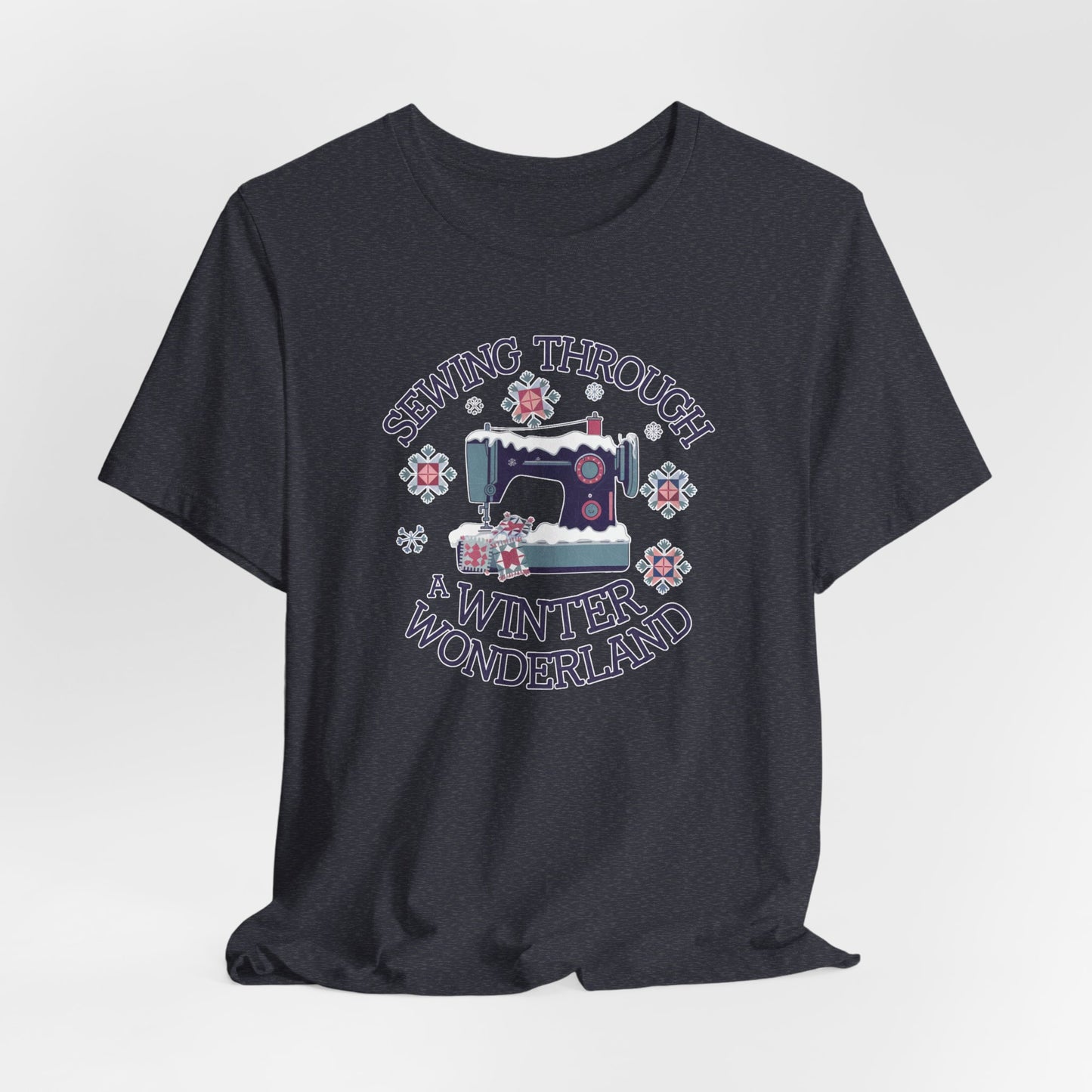 A Heather Navy winter quilting t-shirt featuring  a sewing machine covered in snow, surrounded by quilted snowflakes, with the phrase "Sewing Through a Winter Wonderland" above and below the image.