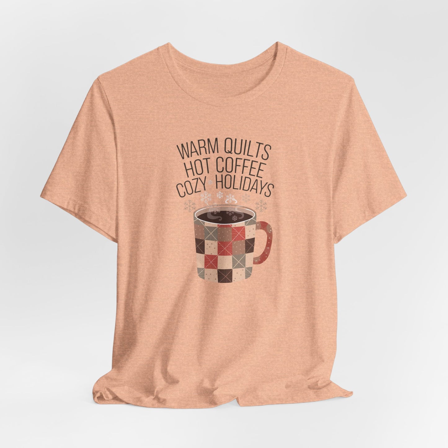 A Heather Peach christmas quilting t-shirt featuring a cozy patchwork quilt-themed coffee mug, surrounded by snowflakes, with the phrase "Warm Quilts, Hot Coffee, Cozy Holidays" above the mug, celebrating the warmth of quilts and coffee during the holiday season.