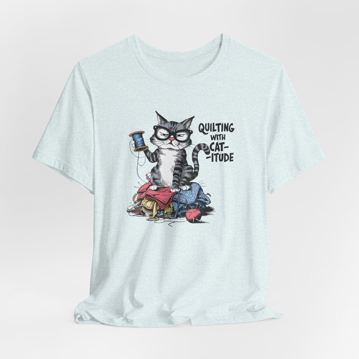 A Heather Ice Blue t-shirt featuring a sassy cartoon cat sitting on a pile of colorful fabric, holding a spool of thread, with the phrase 'Quilting with Cat-itude,' perfect for quilters and cat lovers.
