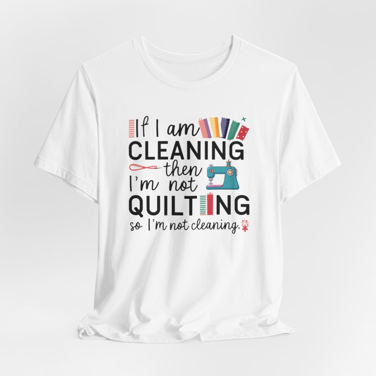 A White funny quilting T-Shirt with the phrase If I am Cleaning then I'm not Quilting in a bold and cheerful font with images of quilting icons and supplies